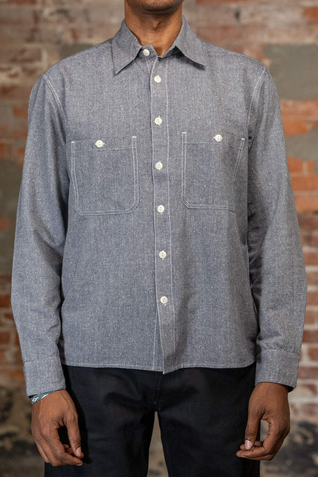 A person shown from the front wearing the 3sixteen Utility Chambray Shirt - Handloom Silk Khadi, while standing in front of a rustic brick wall.