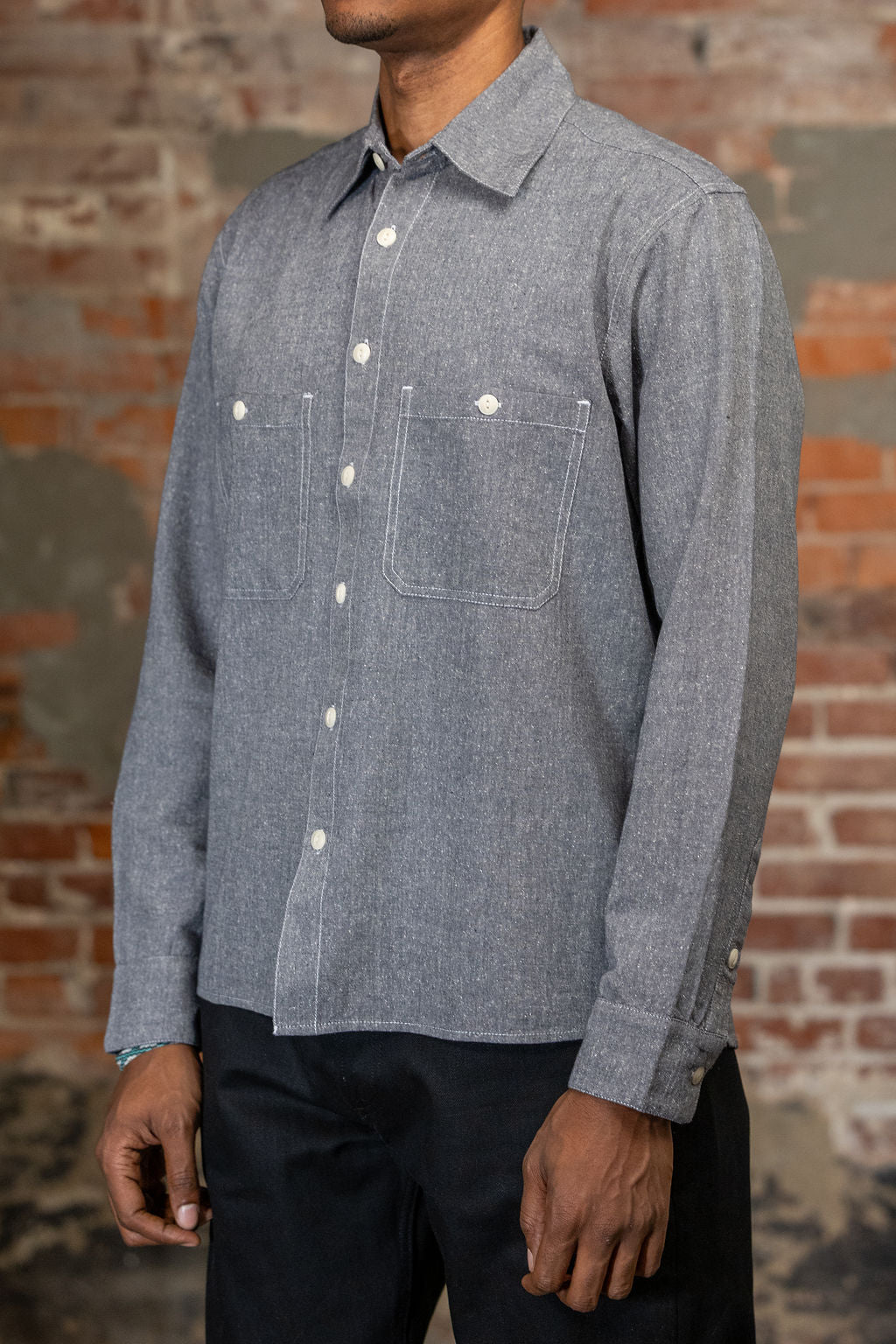 A person shown from the side wearing the 3sixteen Utility Chambray Shirt - Handloom Silk Khadi, while standing in front of a rustic brick wall.