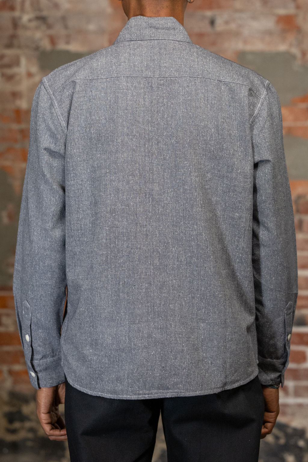 A person shown from the back wearing the 3sixteen Utility Chambray Shirt - Handloom Silk Khadi, while standing in front of a rustic brick wall.