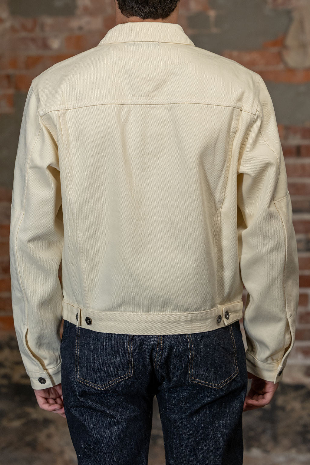 A person viewed from the back wearing the  3sixteen Trucker Jacket - Buttermilk Twill which is a light cream canvas utility jacket and dark blue jeans, standing against a rustic brick wall.