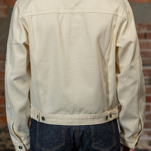 A person viewed from the back wearing the  3sixteen Trucker Jacket - Buttermilk Twill which is a light cream canvas utility jacket and dark blue jeans, standing against a rustic brick wall.