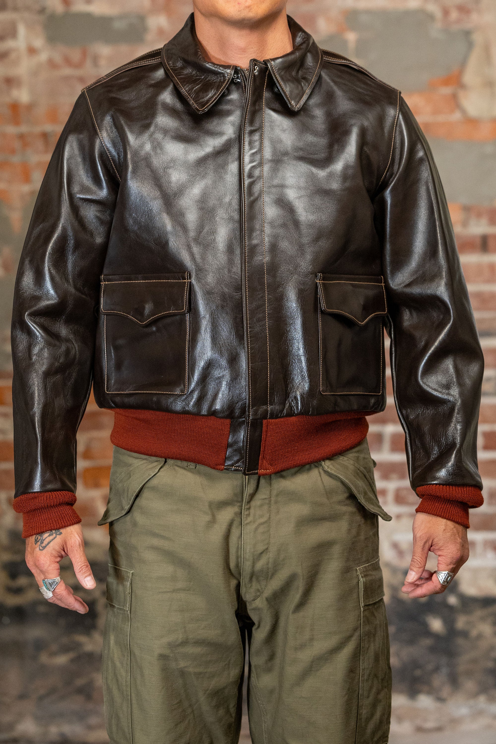 A man viewed from the front wearing  the 
Buzz Rickson's BR80644 Type A-2 “Contract no. w535 AC-21996 Buzz Rickson Clo. Co. - S/Brown, dark brown leather jacket, olive green cargo pants, and a rust-colored sweater, standing against a brick wall.