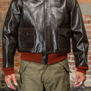 A man viewed from the front wearing  the 
Buzz Rickson's BR80644 Type A-2 “Contract no. w535 AC-21996 Buzz Rickson Clo. Co. - S/Brown, dark brown leather jacket, olive green cargo pants, and a rust-colored sweater, standing against a brick wall.