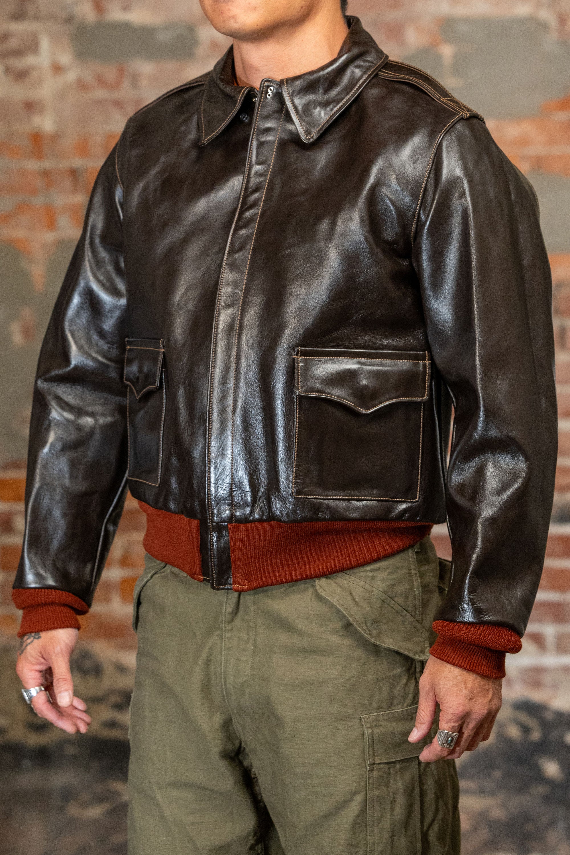 A man viewed from the side wearing  the 
Buzz Rickson's BR80644 Type A-2 “Contract no. w535 AC-21996 Buzz Rickson Clo. Co. - S/Brown, dark brown leather jacket, olive green cargo pants, and a rust-colored sweater, standing against a brick wall.
