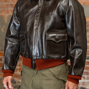 A man viewed from the side wearing  the 
Buzz Rickson's BR80644 Type A-2 “Contract no. w535 AC-21996 Buzz Rickson Clo. Co. - S/Brown, dark brown leather jacket, olive green cargo pants, and a rust-colored sweater, standing against a brick wall.