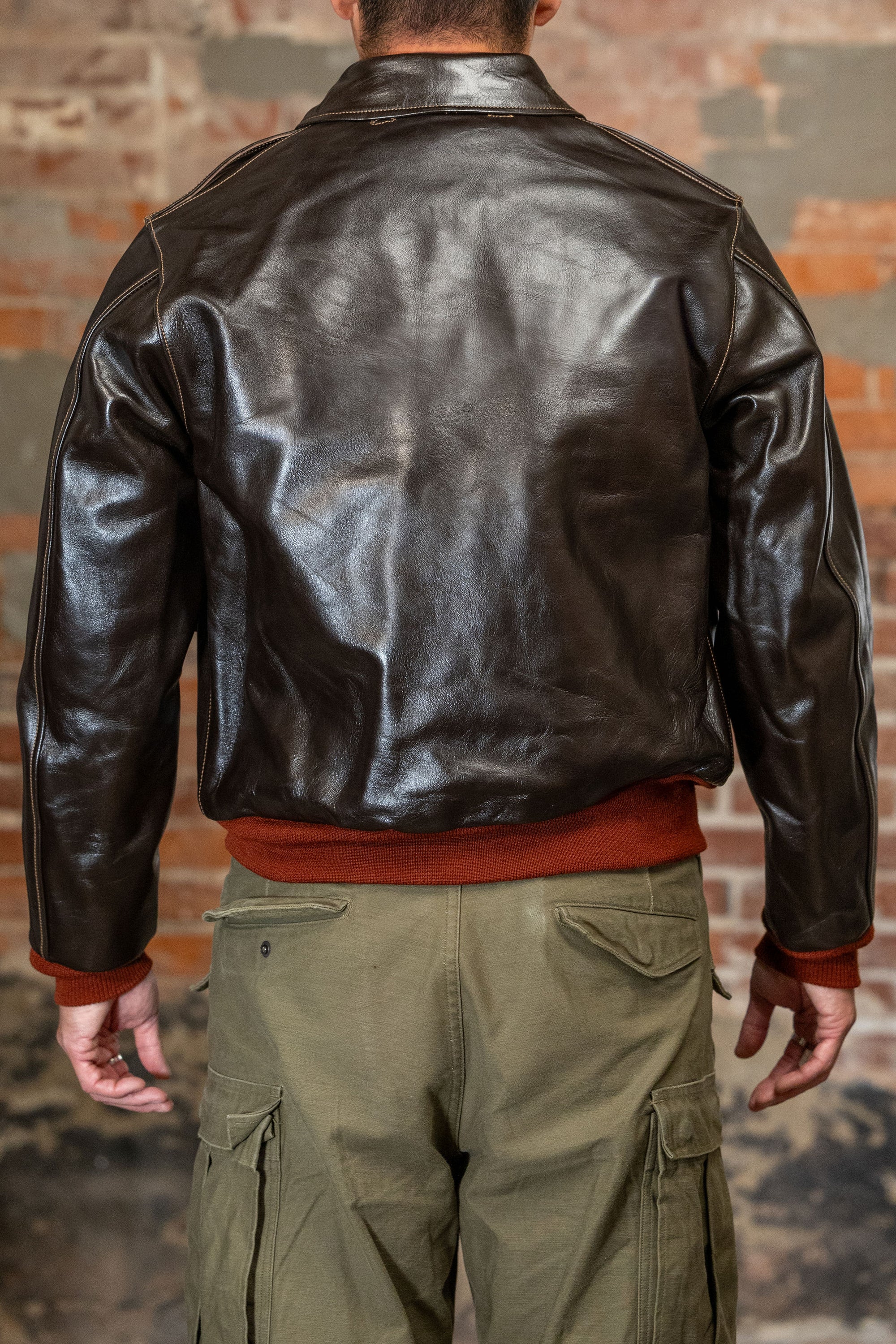 A man viewed from the back wearing  the 
Buzz Rickson's BR80644 Type A-2 “Contract no. w535 AC-21996 Buzz Rickson Clo. Co. - S/Brown, dark brown leather jacket, olive green cargo pants, and a rust-colored sweater, standing against a brick wall.