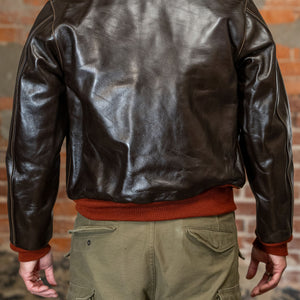 A man viewed from the back wearing  the 
Buzz Rickson's BR80644 Type A-2 “Contract no. w535 AC-21996 Buzz Rickson Clo. Co. - S/Brown, dark brown leather jacket, olive green cargo pants, and a rust-colored sweater, standing against a brick wall.