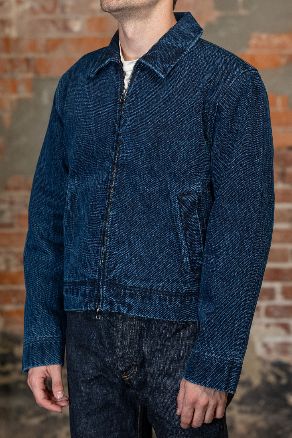 A person viewed from the back wearing 
the 3sixteen Club Jacket - Indigo Kendo Cloth, standing against a rustic brick wall.