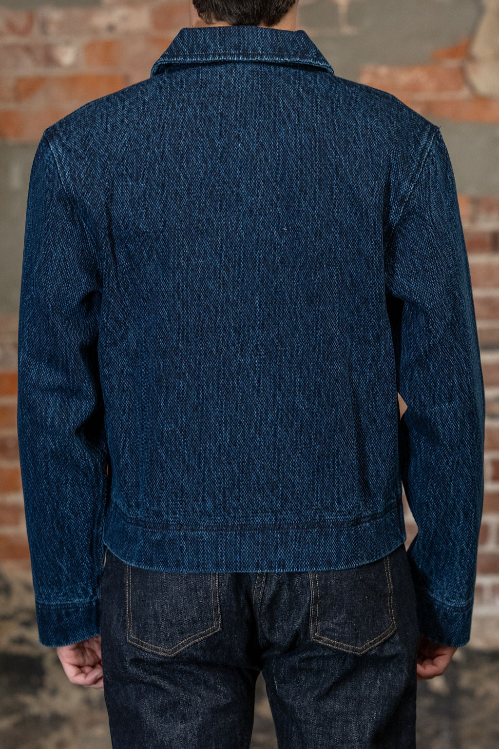 A person viewed from the back wearing 
the 3sixteen Club Jacket - Indigo Kendo Cloth, standing against a rustic brick wall.