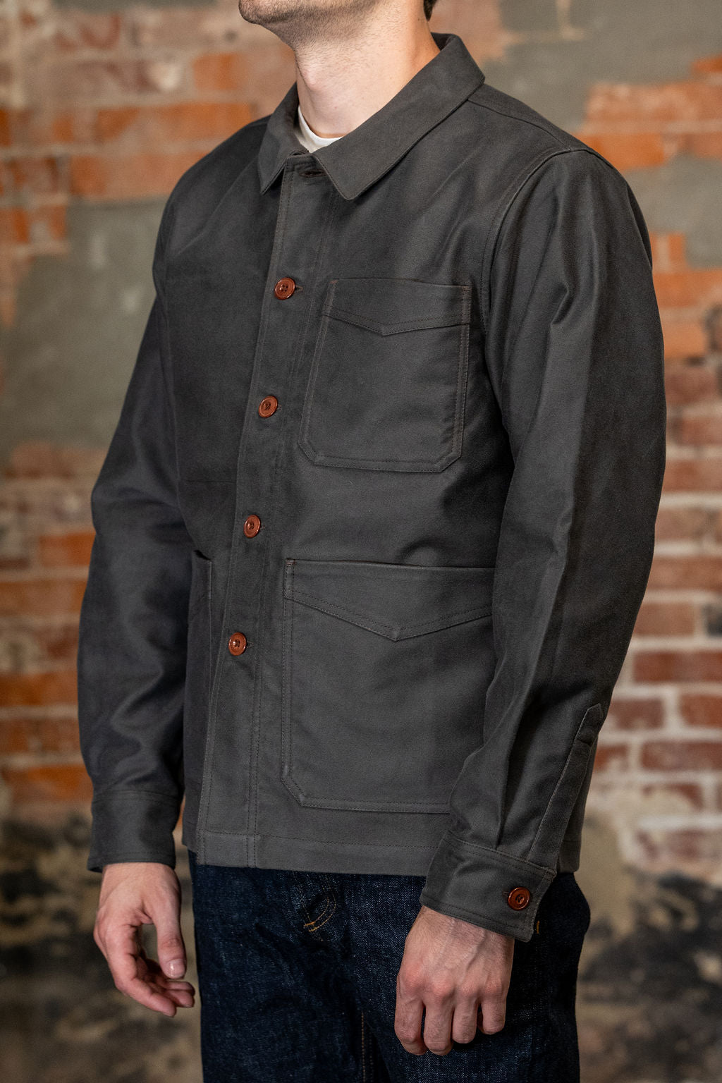 A man viewed from the side wearing
3sixteen French Work Jacket - Grey Moleskin, a dark charcoal gray long-sleeve button-up shirt 