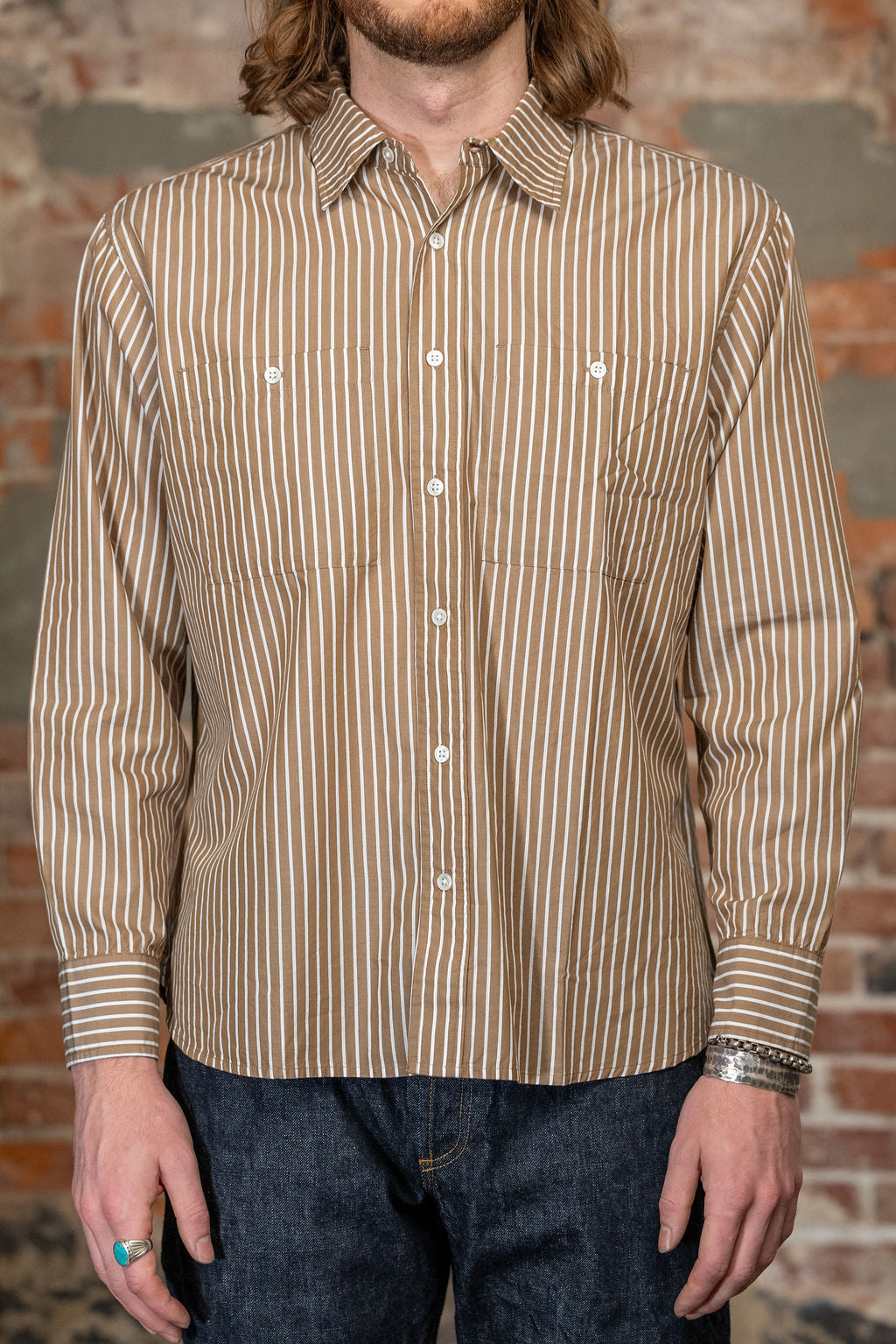 A man viewed from the front wearing the 
3sixteen Commuter Shirt - Brown Ecru Stripe, a beige and brown vertically striped long-sleeve button-down shirt and dark blue jeans, standing against a rustic brick wall.