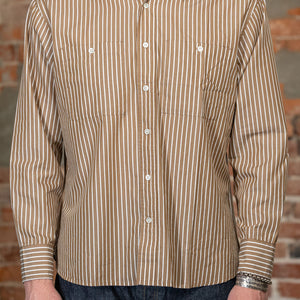 A man viewed from the front wearing the 
3sixteen Commuter Shirt - Brown Ecru Stripe, a beige and brown vertically striped long-sleeve button-down shirt and dark blue jeans, standing against a rustic brick wall.