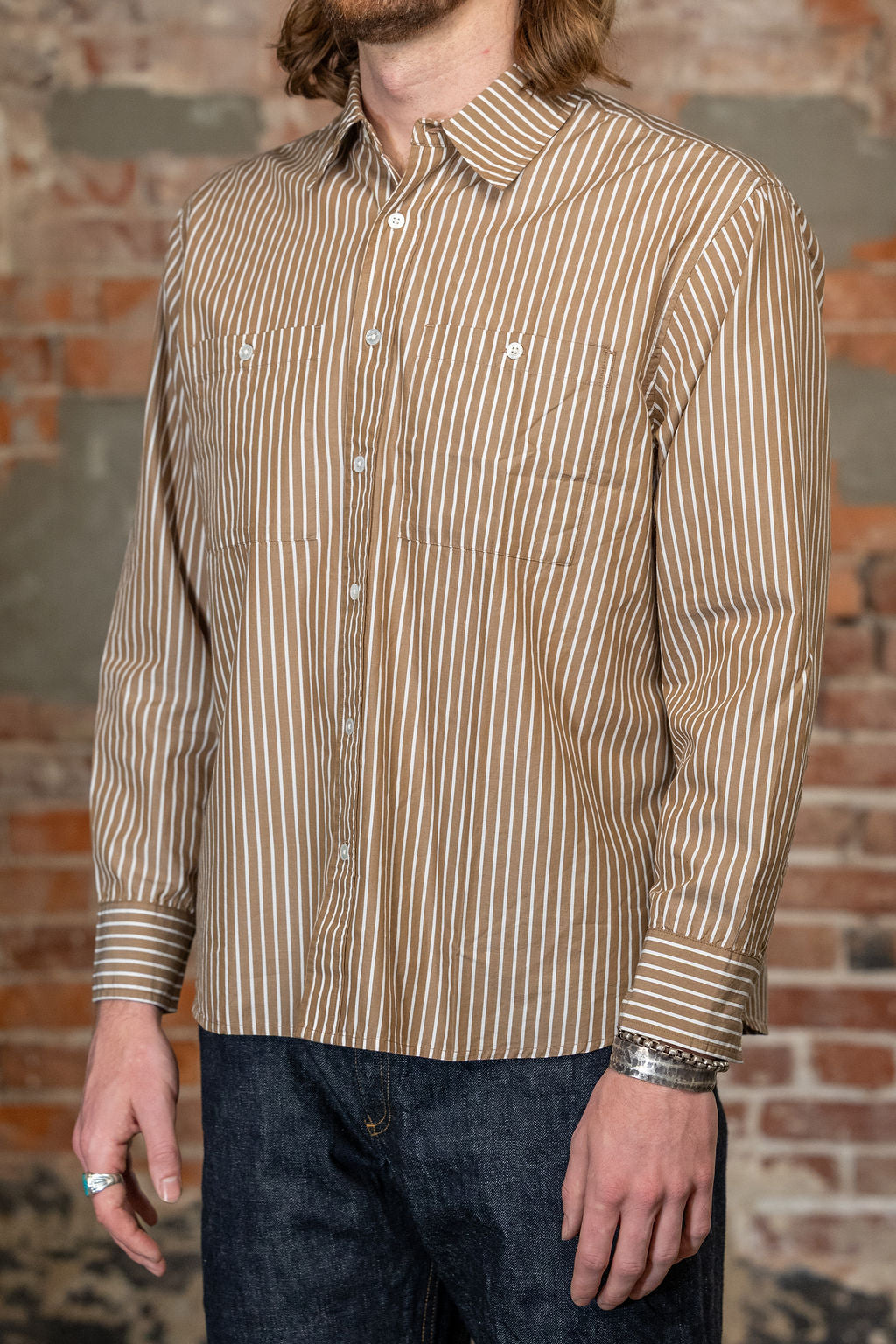 A man viewed from the side wearing the 
3sixteen Commuter Shirt - Brown Ecru Stripe, a beige and brown vertically striped long-sleeve button-down shirt and dark blue jeans, standing against a rustic brick wall.