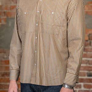 A man viewed from the side wearing the 
3sixteen Commuter Shirt - Brown Ecru Stripe, a beige and brown vertically striped long-sleeve button-down shirt and dark blue jeans, standing against a rustic brick wall.