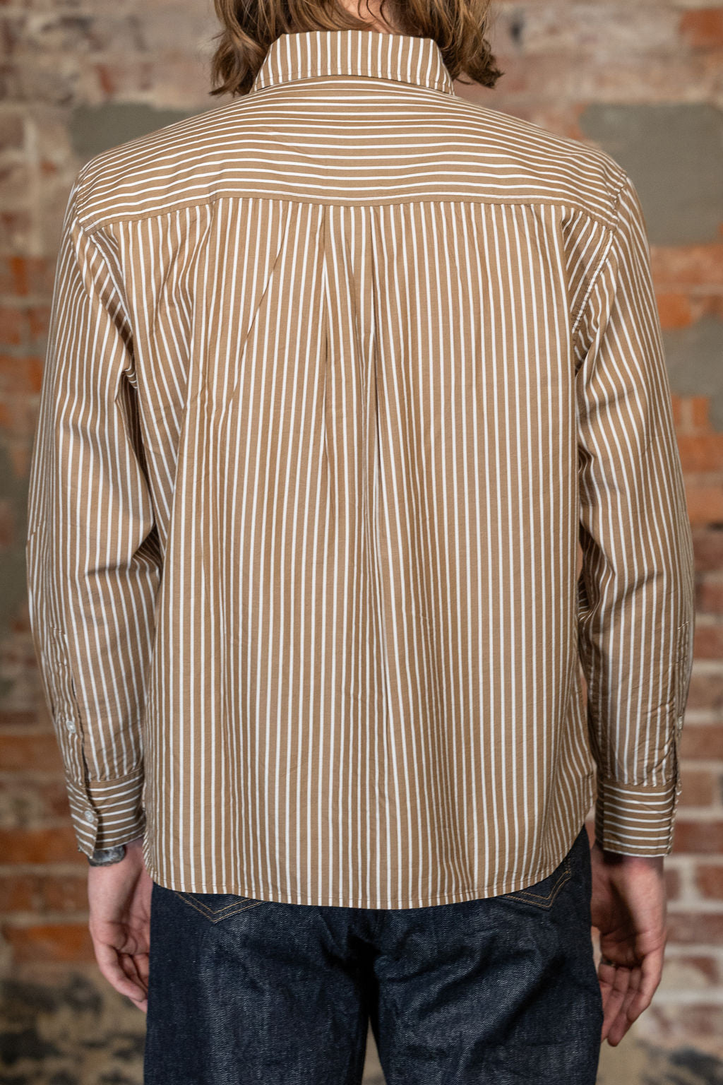 A man viewed from the back wearing the 
3sixteen Commuter Shirt - Brown Ecru Stripe, a beige and brown vertically striped long-sleeve button-down shirt and dark blue jeans, standing against a rustic brick wall.