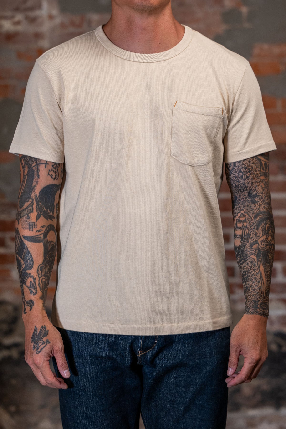 A man wearing the
Freenote Cloth 13oz Pocket T-Shirt in Cream in front of a rustic brick wall.