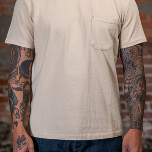 A man wearing the
Freenote Cloth 13oz Pocket T-Shirt in Cream in front of a rustic brick wall.