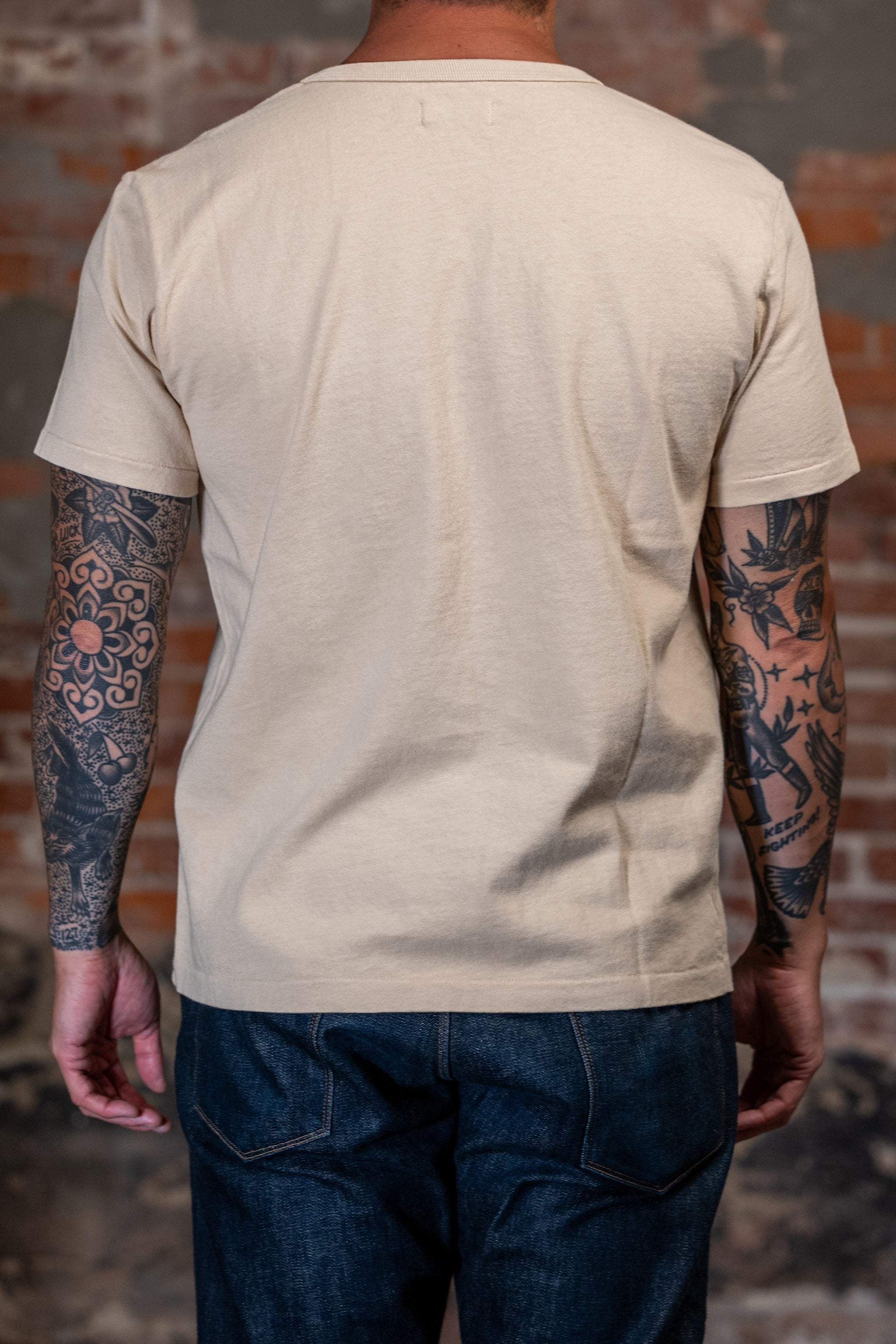 A man viewed from behind wearing the 
Freenote Cloth 13oz Pocket T-Shirt in Cream in front of a rustic brick wall. 