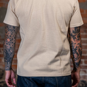 A man viewed from behind wearing the 
Freenote Cloth 13oz Pocket T-Shirt in Cream in front of a rustic brick wall. 